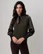 A woman modeling a dark olive green knit sweater vest with long black satin sleeves and a matching black satin bow at the collar, paired with dark brown pants, against a plain light gray studio background.