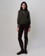 A model stands against a light gray studio background wearing a dark olive green ribbed knit sweater vest over a sheer dark gray long-sleeve blouse, paired with dark brown pants and dark brown knee-high boots.