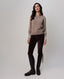 A woman modeling a light taupe knit sweater with dark brown skinny pants and dark brown leather riding boots, standing against a neutral light gray studio background.