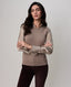 A woman modeling a taupe knit sweater with satin long sleeves and dark brown pants, standing against a light gray studio background.