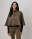 A woman modeling a cropped taupe-brown cape jacket with a front zipper and tassel detail, styled over a dark brown top and matching brown pants, against a neutral light gray background.