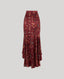 A high-waisted, floor-length mermaid-style skirt in a deep burgundy red, featuring an allover abstract brushstroke pattern in brown, gold, and muted blue, set against a solid light gray background.
