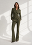 A woman models an olive green double-breasted blazer and matching flared trousers against a white wall with dramatic window shadow patterns.