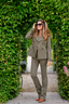 A woman wearing an olive green double-breasted blazer and matching trousers, styled with brown leather shoes and sunglasses, posing in a garden archway made of green hedges.
