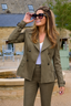 A woman wearing an olive green double-breasted blazer and matching trousers, styled with a white top and black sunglasses, standing outdoors in front of a stone building.