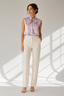 A woman stands in a studio wearing a sleeveless lavender satin blouse with a large bow at the collar and off-white straight-leg trousers, posed against a light gray wall with window shadow patterns.