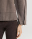 Close-up of a person wearing a taupe knit sweater with ribbed hem and cuffs, paired with dark brown leggings, against a light gray studio background.
