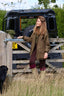 A woman stands outdoors in a rural setting, leaning against a wooden gate in front of a black Land Rover Defender. She is wearing a brown tweed jacket, dark brown trousers, and tall rubber boots.