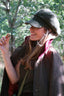A woman outdoors wearing a dark tweed flat cap, a brown wool cape jacket over a pink top, and holding a small acorn in her hand. The background is a soft-focus forest with green foliage.