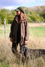 A woman wearing a brown field coat, black top, and green cap stands in a grassy outdoor field with a black dog beside her.