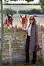A woman posing outdoors in front of a stone wall with two horses behind it. She is wearing a long taupe coat, white top, dark trousers, and brown shoes, holding a dark purple handbag.