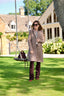 A woman stands on a green lawn in front of a large house, wearing a long taupe-brown coat, dark brown trousers, and sunglasses, with a brown handbag hanging from a swing nearby.