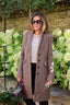 A woman wearing a long taupe-brown double-breasted coat over a cream graphic t-shirt and black trousers, holding a purple handbag, standing outdoors in front of hydrangea bushes and a stone wall.