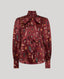 A long-sleeve silk blouse in deep burgundy with an all-over feather print in brown, beige, and black, featuring a high collar and a large bow at the neck, shown against a solid gray background.