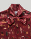A close-up of a collared silk blouse with a large bow at the neck, featuring an all-over feather print in rust red, brown, black, and cream. The brand name 'RALLY WILSON' is visible on the inner neck label.