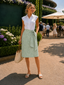 A woman stands outdoors wearing a white sleeveless button-up shirt and a light green textured knee-length skirt tied at the waist, holding a beige tote bag. The background shows a landscaped garden walkway with patio umbrellas and other people in the distance.