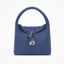 A textured woven blue handbag with a top handle, a matching fabric tie closure, and a round shell-like charm, shown against a plain white background.