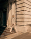 Woman in a beige suit walking along a sunlit street with classical architecture.