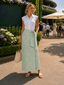 A woman stands outdoors wearing a white sleeveless collared top and a light sage green patterned midi skirt with a side tie, holding a woven straw tote bag. The background shows a garden, patio seating, and other people in a sunny outdoor setting.