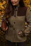 A woman wearing a brown and beige tweed button-front jacket with dark brown suede-like shoulder panels and matching trim, standing outdoors in front of yellow autumn leaves and holding a brown leather handbag.